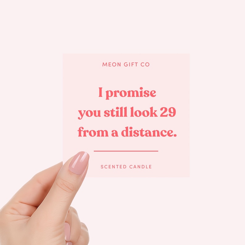 May include: A pink square card held by a hand, with the text "I promise you still look 29 from a distance." The card also includes the text "MEON GIFT CO" and "SCENTED CANDLE".