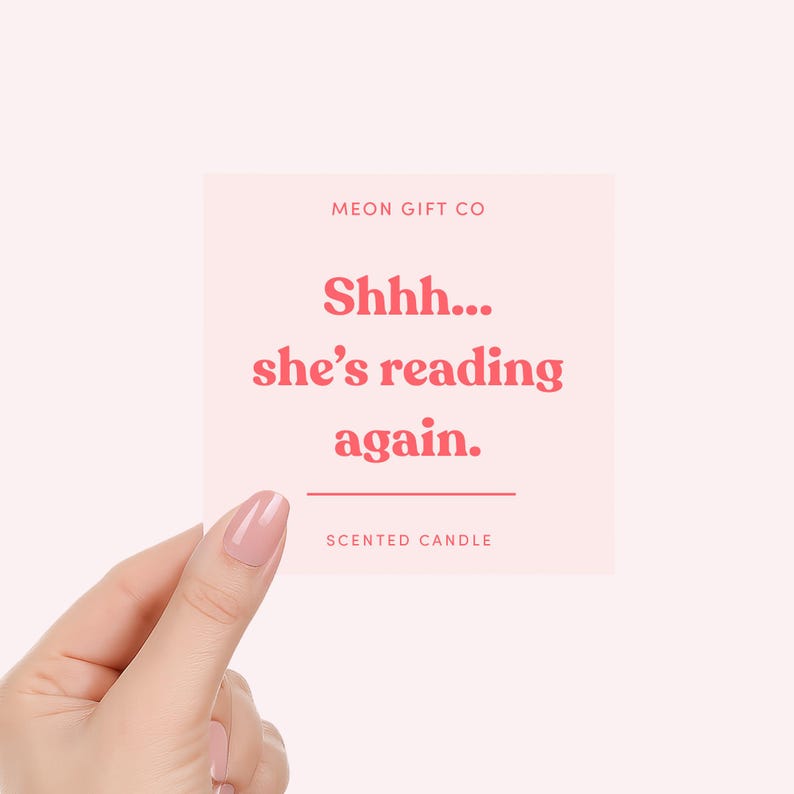 May include: A square, pale pink label with the text "Shhh... she's reading again." in red. The label also includes the text "MEON GIFT CO" and "SCENTED CANDLE" in a smaller font. The label is held by a hand.