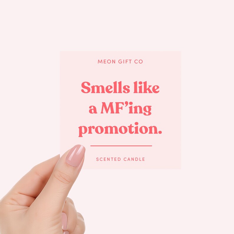 May include: A square, light pink sticker held by a hand, with the text "Smells like a MF'ing promotion." in red. The text "MEON GIFT CO" is at the top, and "SCENTED CANDLE" is at the bottom.