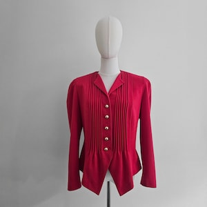 Puede incluir: Una vibrante blazer roja con un corte a medida, con pliegues verticales en la parte delantera y botones dorados. La chaqueta tiene mangas largas y un dobladillo único en punta. La prenda se exhibe en un maniquí.