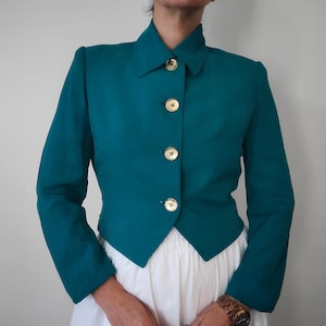 Puede incluir: Una chaqueta azul turquesa recortada con cuello puntiagudo y botones dorados. La chaqueta tiene mangas largas y una silueta ajustada.