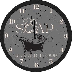 May include: A round wall clock with a black frame and a gray speckled face. The clock features the word "SOAP" and a black bathtub illustration. The phrase "RUB-A-DUB-DUB" is below the tub. The clock hands point to 12 o'clock.