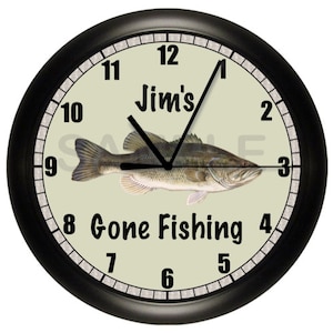 May include: A black framed clock with a green face. The clock has black numbers and hands. The face of the clock has a picture of a bass fish and the text "Jim's Gone Fishing".
