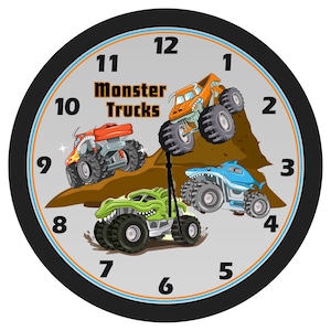 May include: A black and gray clock with a gray face and black numbers. The clock face features three cartoon monster trucks in different colors, driving over a brown dirt mound. The clock face also has the words "Monster Trucks" in black text.