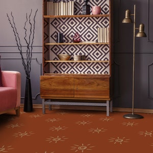 Könnte beinhalten: Ein Wohnzimmer mit einem rosa Sofa, einem Holzbuchregal mit geometrisch gemusterter Tapete, einer schwarzen Vase mit getrockneten Zweigen und einer Stehlampe mit zwei Glühbirnen. Der Boden ist mit einem braunen Teppich mit einem Sonnenmuster bedeckt.