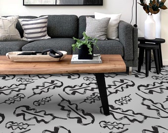 Puede incluir: Una alfombra gris y negra con un patrón repetitivo de hojas de roble estilizadas. La alfombra está en un salón con un sofá gris, una mesa de café de madera y dos mesas auxiliares negras.