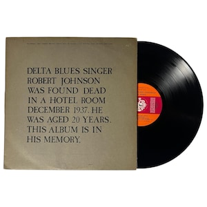 Könnte beinhalten: Ein Vintage-Vinyl-Schallplattenalbum mit einer schwarzen Scheibe und einem beigefarbenen Albumcover. Auf dem Cover steht: "DELTA BLUES SINGER ROBERT JOHNSON WAS FOUND DEAD..." Das Etikett der Schallplatte ist rot und orange.
