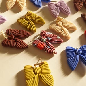Könnte beinhalten: Eine Auswahl an ohrringförmigen Schleifen in verschiedenen Farben, darunter Gelb, Blau, Braun und Lila. Die Ohrringe haben ein strukturiertes, geripptes Design und sind an goldfarbenen Stiften befestigt. Die Ohrringe sind auf einer hellbeige Oberfläche angeordnet.