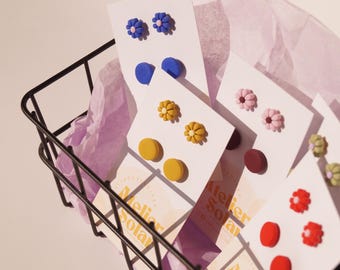 May include: Assortment of colorful polymer clay earrings displayed on white cards with the text "Atelier Solar" printed on them. The earrings feature various shapes, including flowers, circles, and squares.