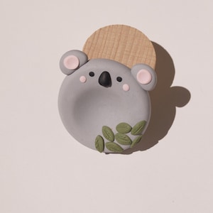 Könnte beinhalten: Ein kreisförmiges, graues Ornament in Koala-Form mit einer schwarzen Nase, kleinen schwarzen Augen, rosa Wangen und rosa Innenohren. Der untere Teil des Koala-Gesichts ist mit kleinen grünen Blättern verziert. Ein hellbrauner Holzhalbkreis befindet sich dahinter.