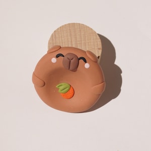 Könnte beinhalten: Eine runde, braune Tonfigur eines Capybaras mit kleinen Ohren, einer dunkelbraunen Nase und rosa Wangen. Das Capybara hält eine kleine orangefarbene Frucht mit einem grünen Blatt in der Mitte. Die Figur ist auf einem runden, hellen Holzsockel befestigt.