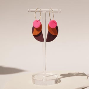 Könnte beinhalten: Ein Paar Ohrhänger mit dünnen goldfarbenen Creolen. Jeder Ohrring besteht aus drei gestapelten geometrischen Formen: einem kleinen leuchtend pinken Kreis, einem mittelgroßen orangefarbenen Halbkreis und einem großen dunkelroten Halbkreis.