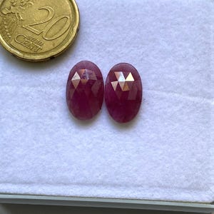 Puede incluir: Dos gemas ovaladas y facetadas, de color rojo intenso, se muestran junto a una moneda de 20 céntimos de euro. Las gemas están sobre una superficie blanca, resaltando sus superficies pulidas y reflejos internos. La moneda sirve como referencia de tamaño.