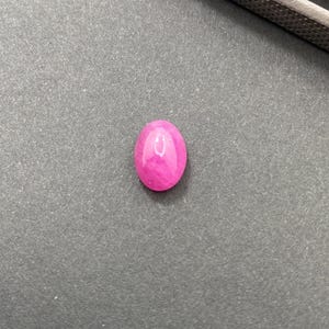 Peut inclure: Une pierre précieuse rose vif polie, de forme ovale. La gemme est lisse et réfléchissante, avec une surface brillante. La pierre est posée sur un fond gris, avec un outil métallique partiellement visible dans le coin supérieur droit.