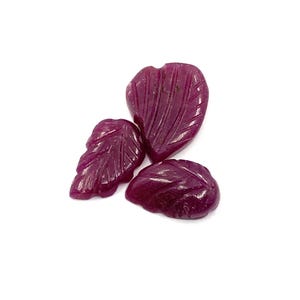 Op de afbeelding: Drie gesneden robijn edelstenen in bladvorm. De edelstenen zijn diep, rijk rood van kleur en hebben gedetailleerde adering. De bladeren variëren in grootte en zijn gerangschikt op een witte achtergrond. Ideaal voor het maken van sieraden.