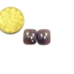 Peut inclure: Deux pierres précieuses taillées carrées, de couleur brun violacé, sont présentées à côté d'une pièce d'or. Les pierres ont une taille en coussin et un aspect légèrement translucide. La pièce est dorée et présente un motif d'étoiles.