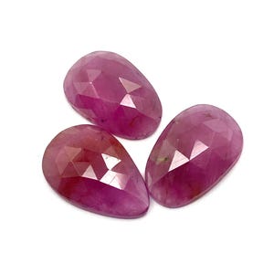 May include: Three faceted, teardrop-shaped gemstones in shades of pink and red. The gems are arranged on a white background, showcasing their polished surfaces and internal facets. The stones have a translucent quality.