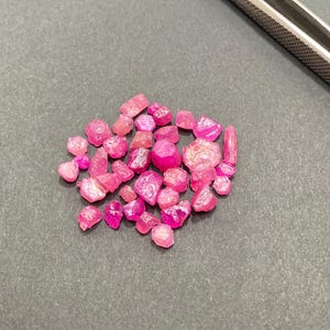 Puede incluir: Un conjunto de gemas de rubí en bruto, sin cortar, de diversas formas y tamaños. Las gemas muestran un vibrante tono rosa a rojo intenso, dispersas sobre una superficie gris oscuro. Se ve un par de pinzas plateadas.