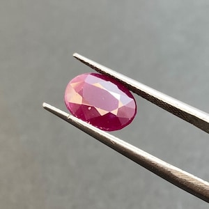May include: A faceted, oval-shaped ruby gemstone held by tweezers. The ruby displays a deep red hue with lighter internal reflections. The background is a solid, neutral gray, providing contrast to the gemstone.