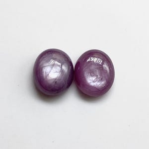 Può includere: Due rubini stellati di forma ovale, tagliati a cabochon, con una tonalità rosso violaceo intenso. Ogni gemma mostra un distinto effetto a stella a sei raggi, noto come asterismo, visibile sulla superficie liscia e lucida al riflesso della luce.