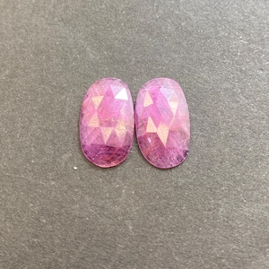 Könnte beinhalten: Zwei ovale, facettierte Edelsteine in Rosa- und Lilatönen. Die Edelsteine haben eine polierte, reflektierende Oberfläche und sind vor einem dunkelgrauen Hintergrund platziert. Die Edelsteine sind wahrscheinlich für die Schmuckherstellung.