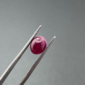 Puede incluir: Un rubí ovalado y pulido, sostenido por unas pinzas plateadas. La gema es de color rojo intenso y tiene una superficie lisa y reflectante. El fondo es gris neutro, resaltando la forma y el color del rubí.