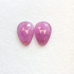 Könnte beinhalten: Zwei facettierte, birnenförmige, rosa Edelsteine. Die Edelsteine haben eine polierte Oberfläche und einen komplexen, geometrischen Schliff. Die Farbe ist ein leuchtendes, mittleres Rosa, mit einigen dunkleren Einschlüssen. Die Edelsteine sind vor weißem Hintergrund.