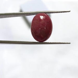 May include: A polished, oval-shaped, deep red gemstone is held by a pair of tweezers. The gem has a smooth, reflective surface, and the color is a rich, saturated shade of red. The tweezers are silver-colored and hold the gem against a white background.