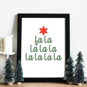 May include: A framed print with a white background and green text that reads "fa la la la la la la la" with a red star at the top. The print is framed in black and is sitting on a wooden surface with two small green artificial Christmas trees.