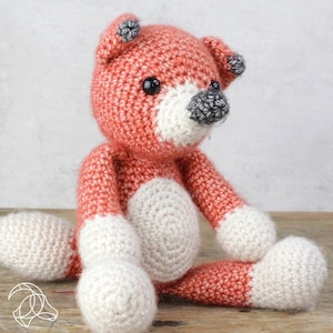 May include: A crocheted fox plush toy with orange and white fur. The fox is sitting with its legs crossed and has black eyes and a gray nose.