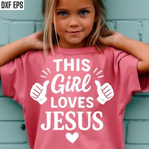 Op de afbeelding: Een roze t-shirt met de witte tekst "THIS GIRL LOVES JESUS". Het ontwerp bevat duimen omhoog en een hart. Het shirt wordt gedragen door een jong meisje met blond haar, tegen een blauwe bakstenen muur.