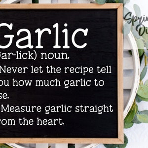 Puede incluir: Una pizarra negra con texto blanco que dice "Garlic (gar-lick) noun. 1. Never let the recipe tell you how much garlic to use. 2. Measure garlic straight from the heart." La pizarra está enmarcada en madera y está rodeada de hojas verdes.