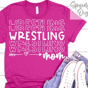 Może przedstawiać: R&oacute;żowa koszulka z białym napisem "Wrestling Mom" z grafiką strzałki i serca.