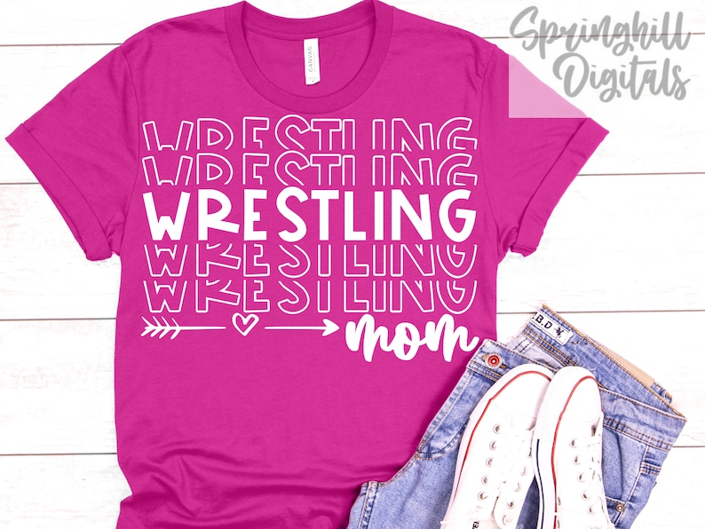 Może przedstawiać: R&oacute;żowa koszulka z białym napisem "Wrestling Mom" z grafiką strzałki i serca.