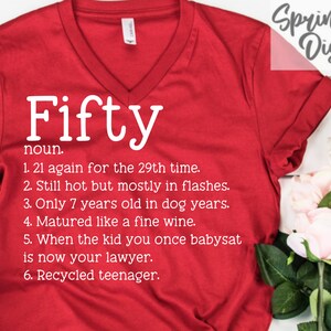 May include: A red V-neck t-shirt with the word "Fifty" in large white letters, followed by a list of humorous definitions. The text "Springhill Digitals" is in the upper right corner. The shirt is displayed on a white surface with flowers.