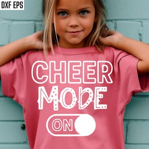 May include: A young girl wearing a pink t-shirt with the words "CHEER MODE ON" in white block letters. The shirt has a switch graphic with the word "ON" inside. The background is a light blue brick wall.