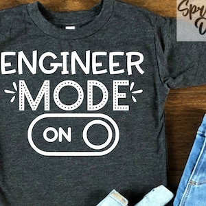 May include: A dark gray t-shirt with white text that reads "Engineer Mode On" with a white toggle switch graphic.