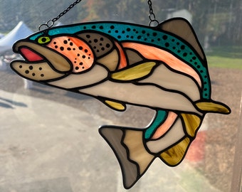 May include: A stained glass art piece depicting a trout fish with a teal, pink, and brown color scheme. The fish is suspended from a black chain.