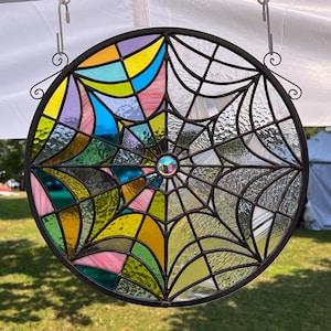 May include: A round stained glass window with a spiderweb design. The left side features colorful geometric shapes in yellow, pink, blue, and green. The right side has a clear web pattern. A decorative metal frame surrounds the glass.