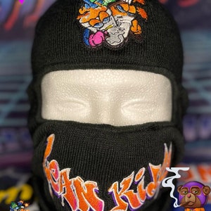 Puede incluir: Máscara de esquí negra con un gráfico "Lean Kids" naranja y morado. La máscara de esquí tiene un gorro negro con un personaje de dibujos animados bordado en la parte superior.