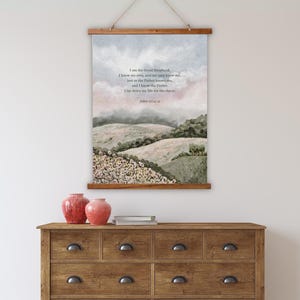 Puede incluir: Un tapiz de pared de madera con una pintura de paisaje y texto de Juan 10:14-15. La pintura representa colinas onduladas bajo un cielo nublado. El tapiz se muestra encima de una cómoda de madera con nueve cajones.