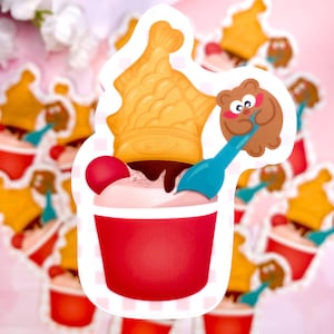 Könnte beinhalten: Ein roter und weißer Eisbecher mit Schokoladensoße, einem gelben, fischförmigen Waffelhörnchen, einem braunen Bären, der einen blauen Löffel hält, und einer roten Kirsche oben drauf.