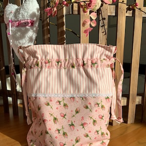 Könnte beinhalten: Eine rosa-weiße gestreifte Kordeltasche mit einem rosa Blumendruck auf dem Boden. Die Tasche hat einen weißen Spitzenbesatz und ist aus Baumwollstoff gefertigt.
