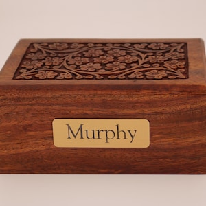 May include: A rectangular wooden box with a floral carved design on the lid. The box is a warm brown color and has a gold nameplate that reads "Murphy".