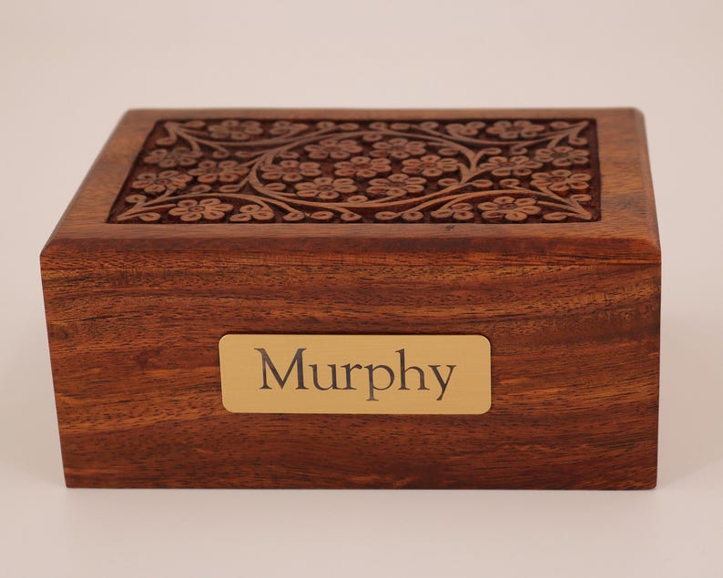 May include: A rectangular wooden box with a floral carved design on the lid. The box is a warm brown color and has a gold nameplate that reads "Murphy".