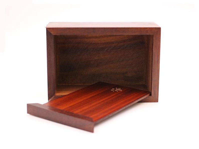 May include: A rectangular wooden box with a sliding tray. The box and tray are made of dark brown wood with a visible wood grain. The tray is partially extended, revealing a gold-colored design.