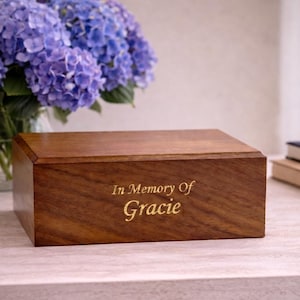 May include: A rectangular wooden box with a smooth, dark brown finish and a beveled lid. The front panel features the gold-colored engraved text In Memory Of Gracie, centered in a serif font. The box has a visible wood grain texture throughout.
