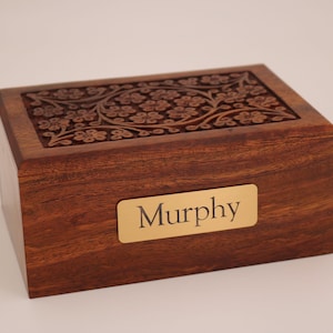 May include: A rectangular wooden box with a floral carved design on the lid. The box is a rich brown color with a gold nameplate that reads "Murphy". The box is likely used for keepsakes or as a decorative item.