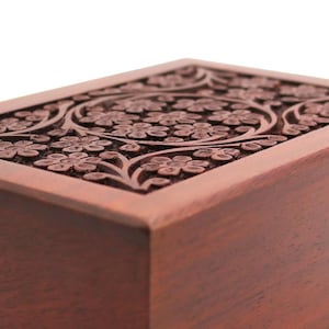 May include: A rectangular wooden box with a detailed floral carving on the lid. The box is a warm brown color, and the carving features a repeating pattern of flowers and leaves. The wood grain is visible, adding to the natural aesthetic.