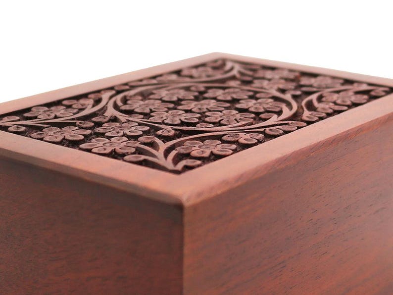 May include: A rectangular wooden box with a detailed floral carving on the lid. The box is a warm brown color, and the carving features a repeating pattern of flowers and leaves. The wood grain is visible, adding to the natural aesthetic.